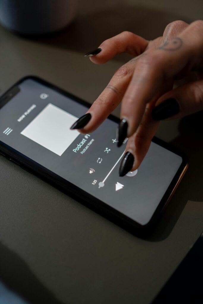 A hand with tattoos uses a smartphone touchscreen to play a podcast indoors.